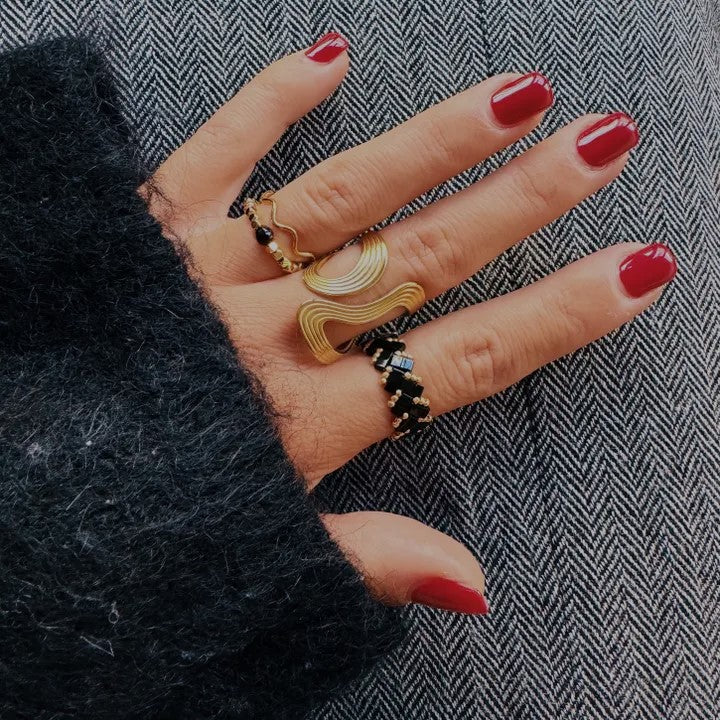 Eine hand mit rot lackierten Fingernägeln. An den Fingern sind unterschiedliche Statement ringe in gold und schwarztönen aus robustem Edelstahl. Die Hand liegt auf einem dunklen untergrund.