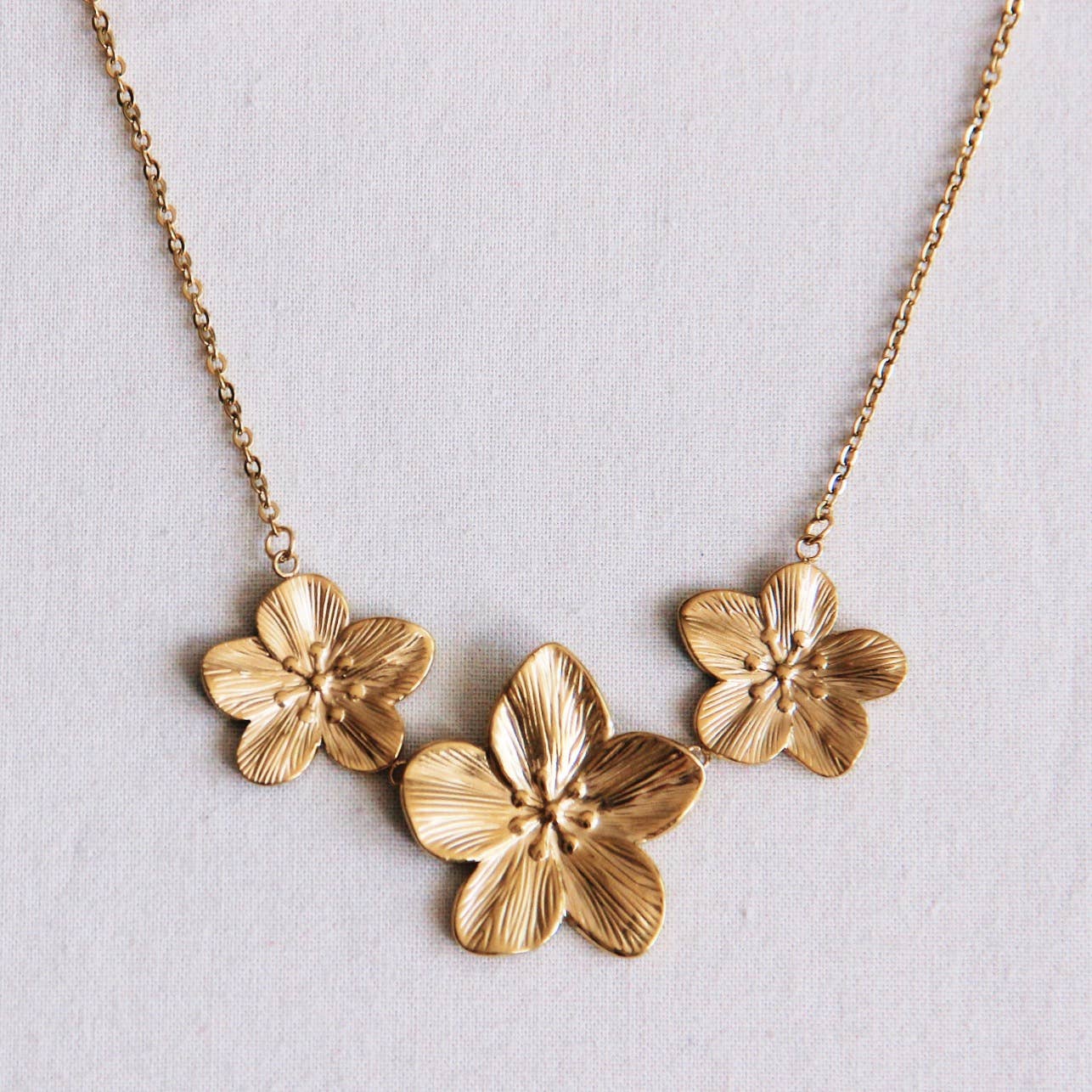 Filigrane Kette mit drei Statement Blumen. In goldene farbe aus robustem Edelstahl, perfekt um aufzufallen und für einen schicken, trendy look. Die Kette liegt auf einem hellen untergrund.