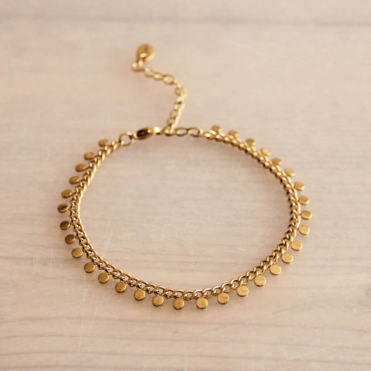 Ein Armband aus Edelstahl liegt auf einem beigen untergrund. An edm Armband hängen goldenene Münzen.