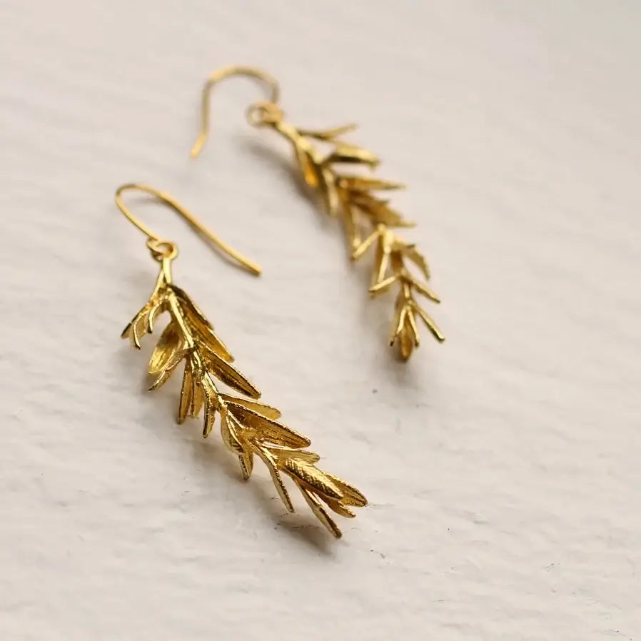 Zwei goldfarbene Messing Ohrringe mit Rosmarin Zweigen als Anhänger liegen auf weißem Papier. Für einen natürlichen und eleganten Look.