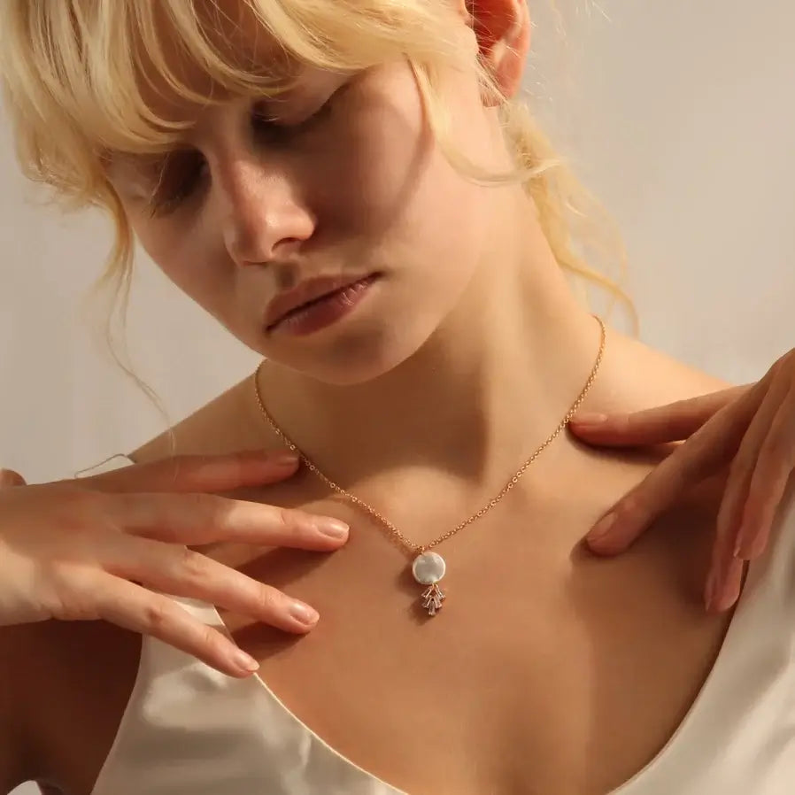 Eine Frau von vorne. Sie trägt eine filigrane, goldfarbene Kette mit einem Perlenanhänger. Unter der Perle sind geometrisch angeordnete Kristalle in einer goldfarbenen Fassung.