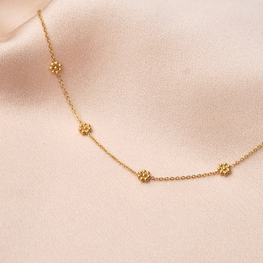 Zarte Edelstahlkette mit goldenen Blumenanhängern auf sanftem Untergrund.