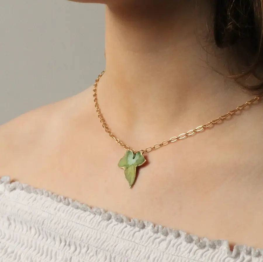 Das Dekoltee einer Frau, die eine feine, goldfarbene Kette mit einem olivgrünen Efeublatt Anhänger trägt. Der Anhänger ist ca 2cm lang und ist perfekt für alle, die Schmuck mit Naturmotiven mögen.