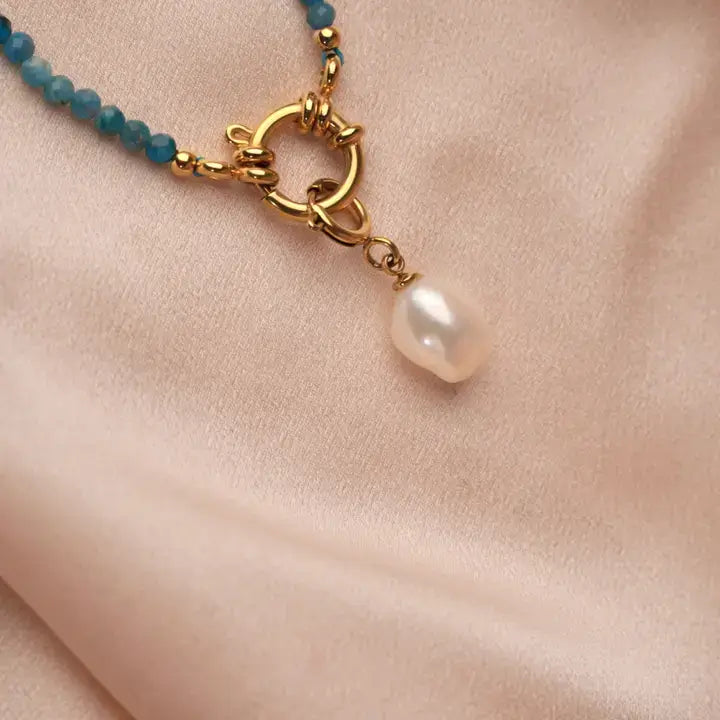Kette mit blauen Natursteinen und goldfarbenem Edelstahl Anhänger mit Perle, elegant auf satiniertem Stoff.