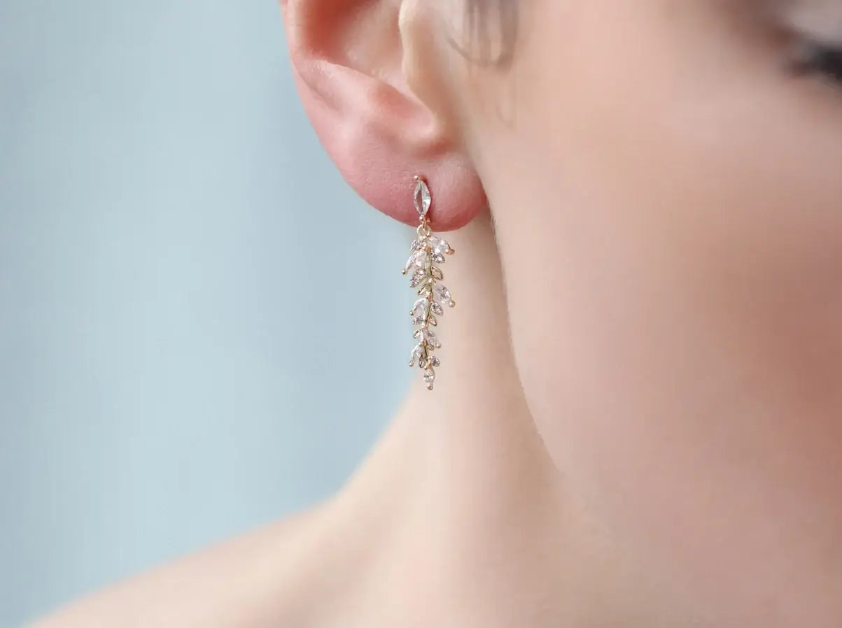 Das Ohr und Gesicht einer Frau von vorne. Am Ohr hängt ein Ohrring in Form einer Feder oder eines Farn Blattes. Der Ohrring ist goldfarben und auf den Blättern sind viele, kleine diamantartige, glitzernde Steine. 