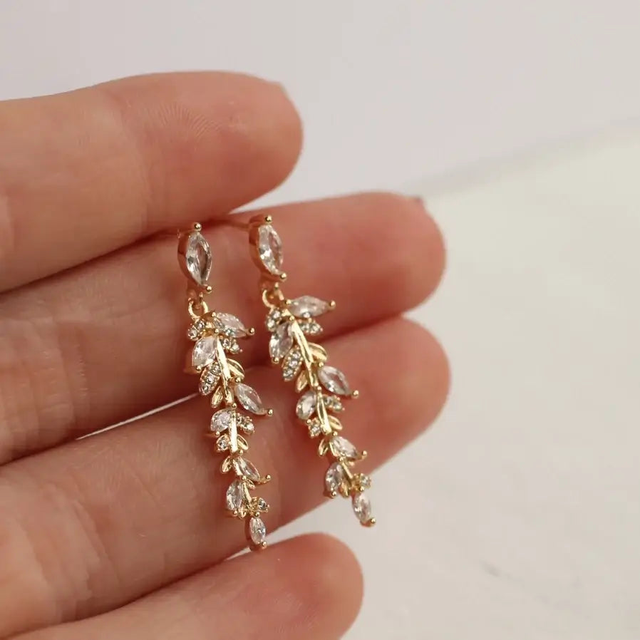 Eine Hand hält zwei längliche Ohrringe mit Kristall und Glasstein details in Form von Federn oder Farnblättern für einen edlen natürlichen Stil. Perfekt für feierliche Anlässse und elegante Looks.