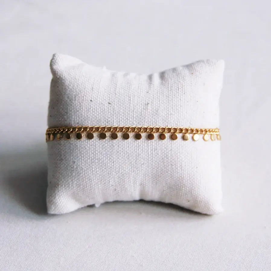 Ein goldfarbenes Armband mit vielen kleinen, runden, goldfarbenen Anhängern ist um ein kleinen Kissen angebracht. Das Armband ist perfekt für einen dezenten, femininen Look.