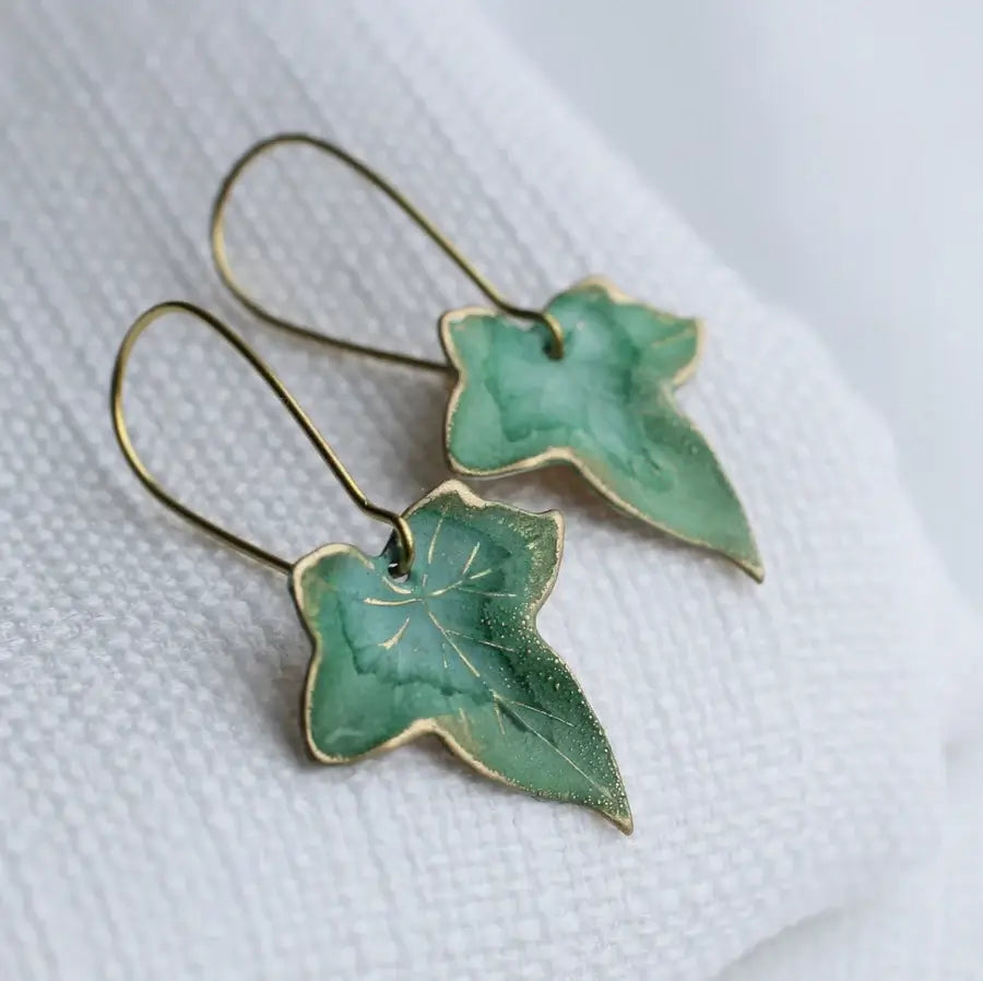 Zwei goldfarbene Messing Ohrringe mit grünen, ca 2cm großen, Efeublatt Anhängern die auf weißem Papier liegen. Für alle die Schmuck mit Natur Optik lieben.